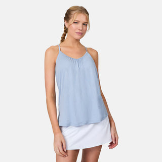 A blonde woman with a braid wears the Free Country Women's Microtech® Chill B Cool V-Neck Cami Top with Built-In Bra in light blue and a white skirt, standing against a plain background and smiling slightly at the camera.