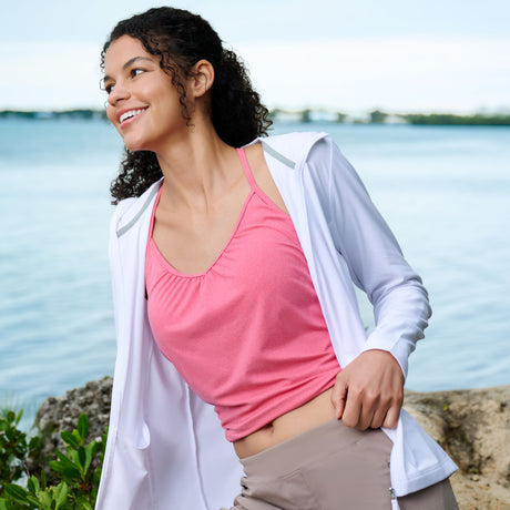A woman with curly hair smiles by the water, enjoying a sunny day in lush greenery and blue sky. She wears a Free Country Women’s SunFree Full Zip UPF Sunshirt in white over a pink top, featuring moisture-wicking fabric for all-day comfort.