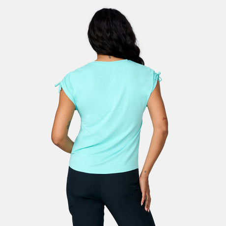 A woman faces away against a white background, wearing the Free Country Women's Microtech® Chill Dolman Sleeve Top in light blue with gathered shoulders and dark pants.