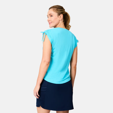 A woman with light brown hair in a ponytail models the Free Country Women's Microtech® Chill Dolman Sleeve Top in light blue with a navy skirt, looking over her shoulder against a plain white background.
