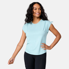 A woman with long wavy hair wears a light blue Free Country Women's Microtech® Chill Dolman Sleeve Top and black pants, smiling to the side with one hand on her hip. The quick-drying top offers UPF 50+ sun protection. The background is plain white.