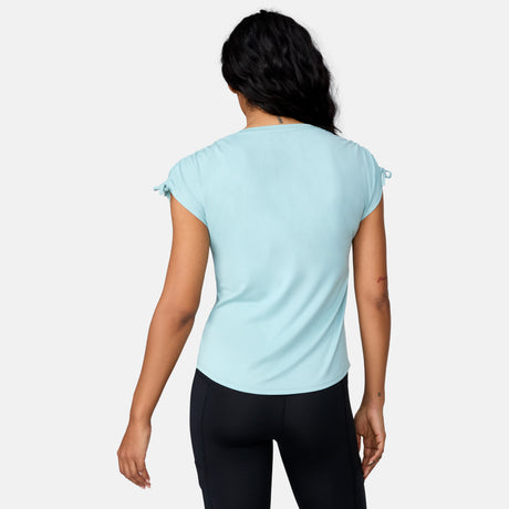 A woman stands with her back to the camera, wearing a light blue Free Country Women's Microtech® Chill Dolman Sleeve Top with UPF 50+ protection and black leggings, set against a plain white background.