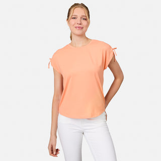 A woman with blonde hair pulled back stands against a light background, smiling softly in a peach Free Country Women's Microtech® Chill Dolman Sleeve Top with small sleeve bows and white pants.