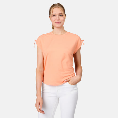 A woman with blonde hair tied back smiles slightly, posing against a white background in the Free Country Women's Microtech® Chill Dolman Sleeve Top in light peach, featuring small sleeve bows, paired with white pants.