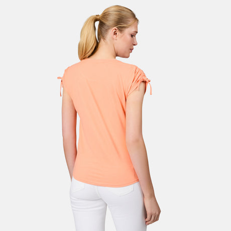A woman with blonde hair in a ponytail is shown from the back, wearing the Free Country Women's Microtech® Chill Dolman Sleeve Top in peach and white pants, standing against a plain light background.