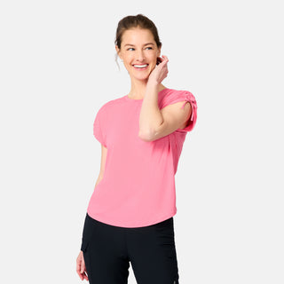 A smiling woman with light brown hair in a ponytail wears a pink Women's Microtech® Chill Dolman Sleeve Top by Free Country and black pants. She stands against a plain light background, touching her face and looking slightly to the side.