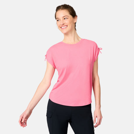 A smiling woman wears the Free Country Women's Microtech® Chill Dolman Sleeve Top in pink with small sleeve bows and black pants, standing against a plain white background.