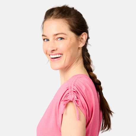 A smiling woman with light skin and a braided brown hairstyle wears the Free Country Women's Microtech® Chill Dolman Sleeve Top in pink, standing against a plain light background.