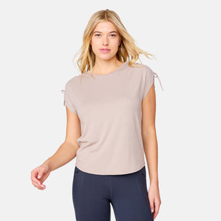 A woman with light blonde hair wears the Free Country Women's Microtech® Chill Dolman Sleeve Top in beige and dark leggings, posing against a plain white background and smiling slightly at the camera.