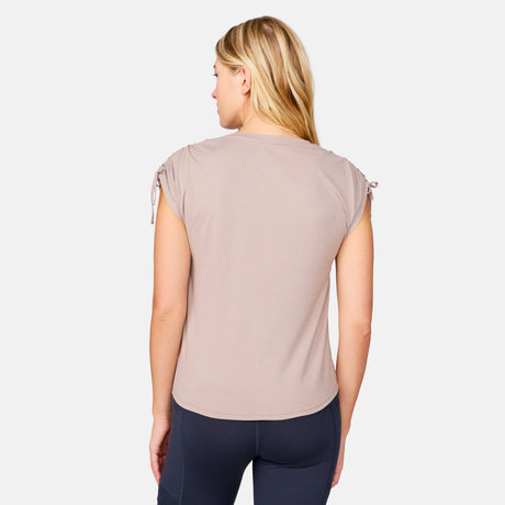 A blonde woman is seen from behind wearing the Free Country Women's Microtech® Chill Dolman Sleeve Top in taupe with drawstring sleeves and dark leggings, standing out against a plain, light-colored background.