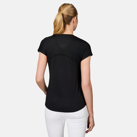 A woman with blonde hair in a ponytail is seen from behind, wearing a black Free Country Women's Microtech® Chill B Cool Tee and white pants, standing against a plain light gray background.