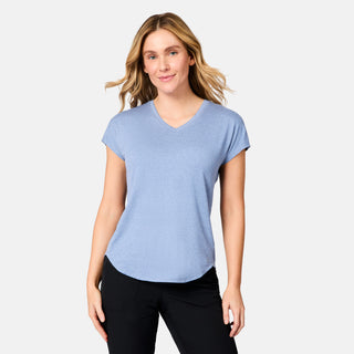A woman with blonde hair smiles softly at the camera, wearing the Free Country Women's Microtech® Chill B Cool Tee in light blue and black pants, standing against a plain light gray background.