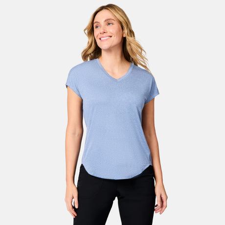 Smiling woman with wavy blonde hair wearing the Free Country Women's Microtech® Chill B Cool Tee in light blue and black pants, standing against a plain light gray background.