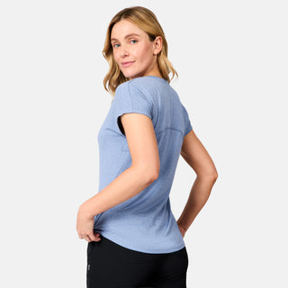A blonde woman stands slightly to the side, looking over her shoulder in a neutral pose, wearing the Free Country Women's Microtech® Chill B Cool Tee in light blue and black pants against a plain light background.