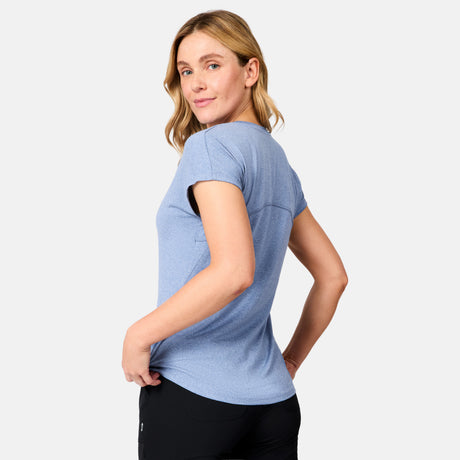 A blonde woman stands slightly to the side, looking over her shoulder in a neutral pose, wearing the Free Country Women's Microtech® Chill B Cool Tee in light blue and black pants against a plain light background.