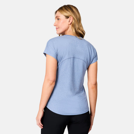 A blonde woman faces away, looking over her left shoulder. She wears the Free Country Women's Microtech® Chill B Cool Tee in light blue with black pants against a plain light background.