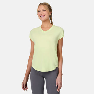 A woman with blonde braided hair wears the Free Country Women's Microtech® Chill B Cool Tee in light yellow and gray leggings, smiling while standing against a plain white background.