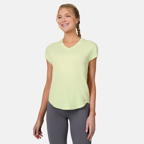 A woman with blonde braided hair wears the Free Country Women's Microtech® Chill B Cool Tee in light yellow and gray leggings, smiling while standing against a plain white background.