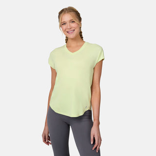A blonde-haired woman in braids smiles while wearing the Free Country Women's Microtech® Chill B Cool Tee in light yellow and gray leggings, standing against a plain white background.