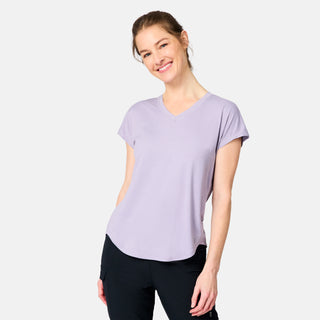 A woman with light brown hair tied back smiles with one hand in her pocket, wearing the Free Country Women's Microtech® Chill B Cool Tee in light purple and black pants against a plain light gray background.
