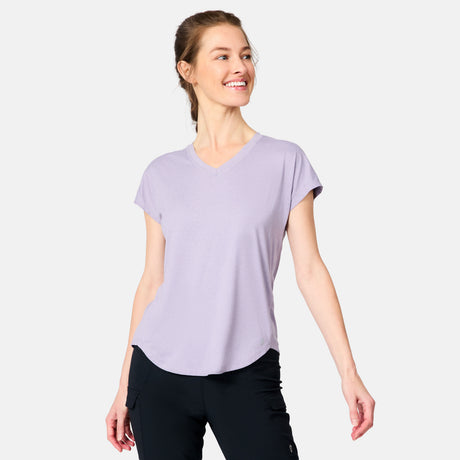 A woman smiles with her hair tied back, wearing the Free Country Women's Microtech® Chill B Cool Tee in light purple and black pants, standing against a plain light background with arms relaxed at her sides.