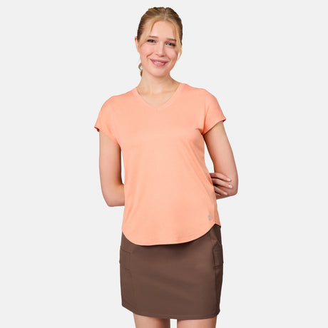 A woman stands against a light background, smiling. She wears a Free Country Women's Microtech® Chill B Cool Tee in peach and a brown skirt with pockets, with one hand resting on her arm.
