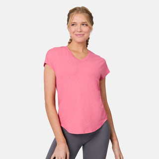 A blonde woman with braided hair smiles and looks to the side, wearing the Free Country Women's Microtech® Chill B Cool Tee in pink and gray leggings, standing against a plain white background.