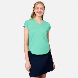 A smiling woman with long blonde hair wears the Free Country Women's Microtech® Chill B Cool Tee in light green and a navy blue skirt, standing against a plain white background.