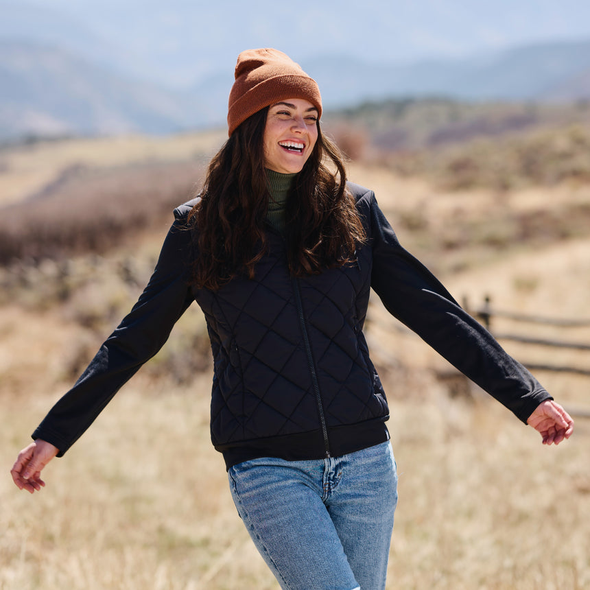Woman in a quilted jacket and beanie standing in a field with mountains in the background