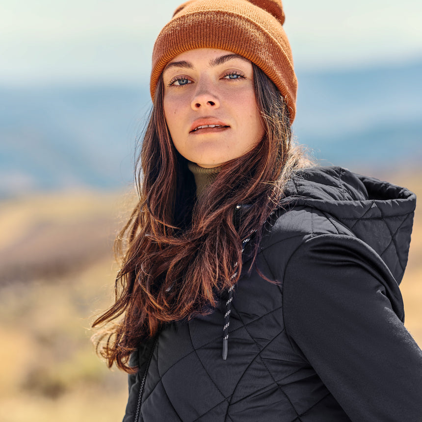 Woman wearing a black quilted jacket and orange beanie outdoors with a blurred natural background