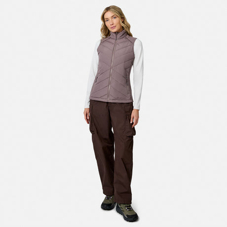 A woman wears a mauve Free Country Women’s Quilted Hybrid Vest over a white long-sleeved shirt, brown cargo pants, and hiking shoes.