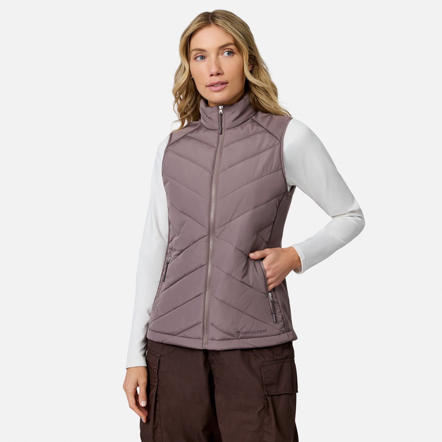 A woman wearing a mauve, lightweight Free Country Women’s Quilted Hybrid Vest over a white long-sleeve shirt and brown cargo pants.