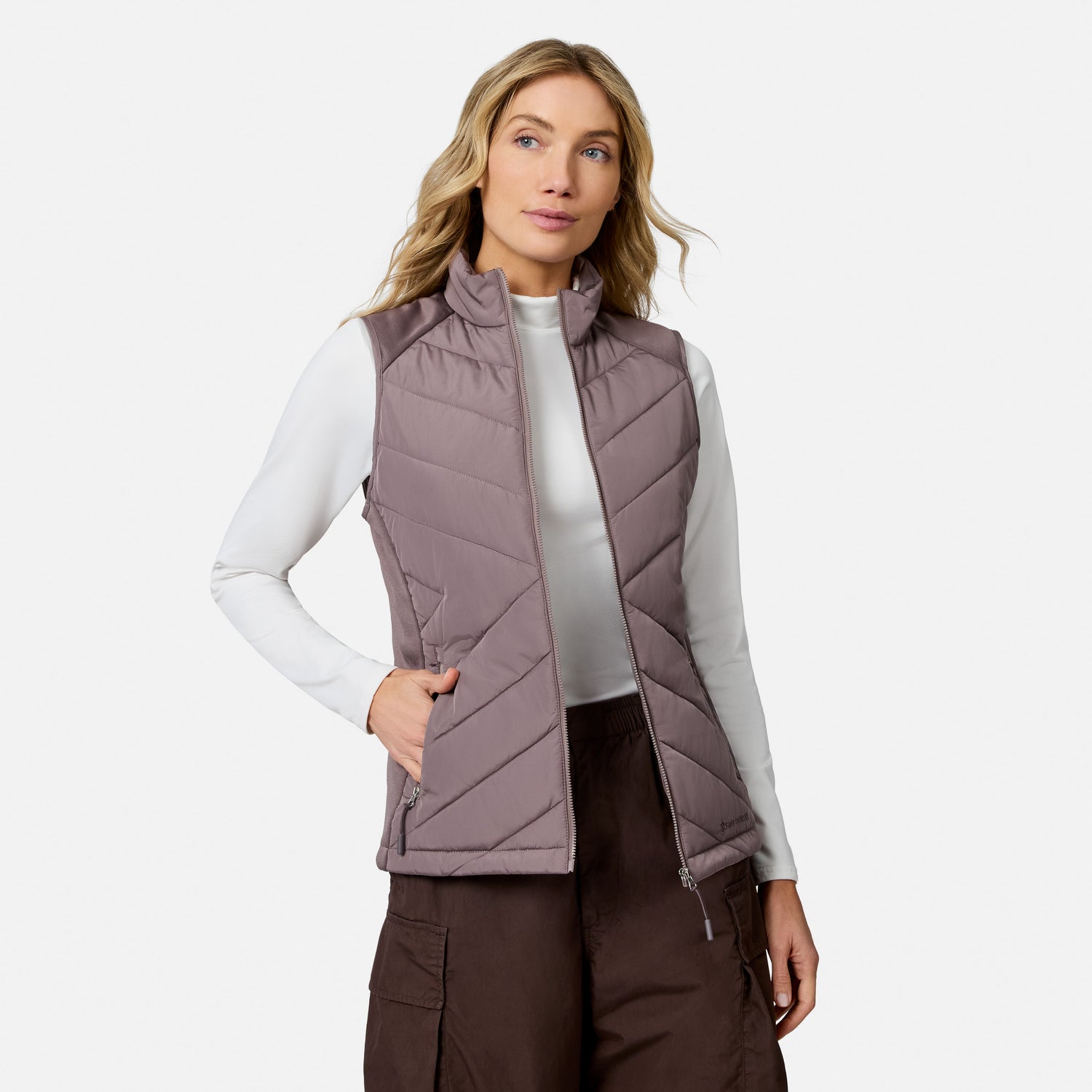 A person wears a lightweight mauve Free Country Women’s Quilted Hybrid Vest over a white long-sleeved shirt and dark brown cargo pants.