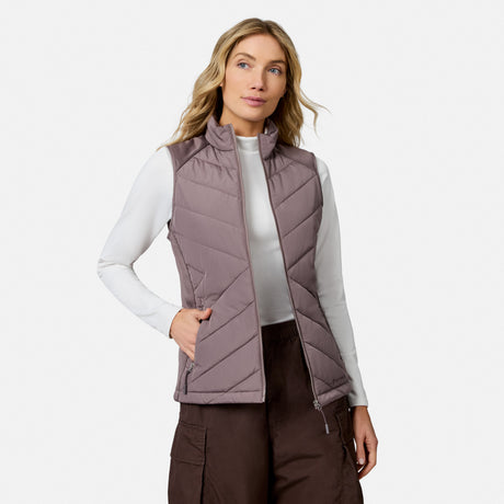 A person wears a lightweight mauve Free Country Women’s Quilted Hybrid Vest over a white long-sleeved shirt and dark brown cargo pants.