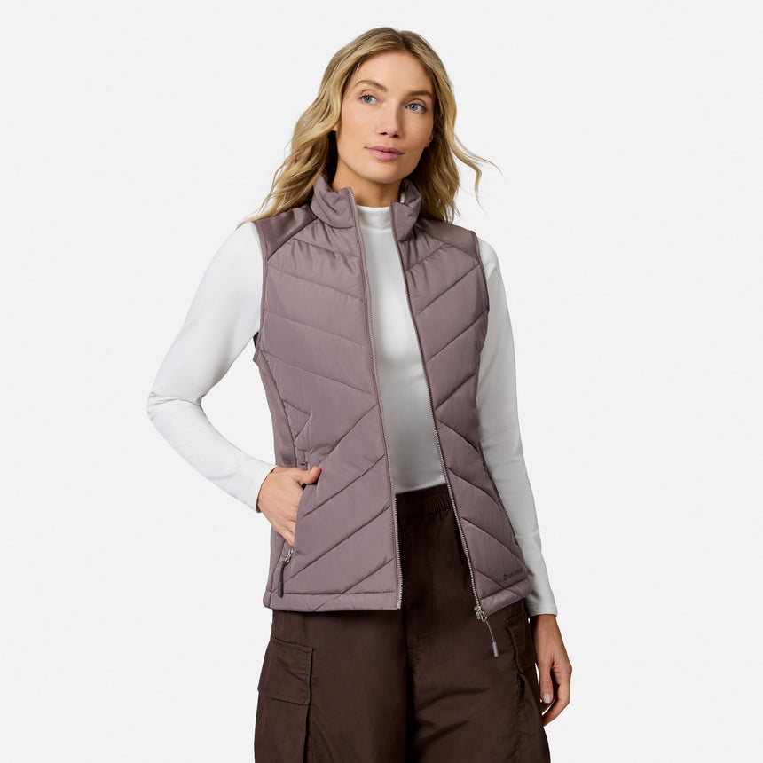 A person wears a lightweight mauve Free Country Women’s Quilted Hybrid Vest over a white long-sleeved shirt and dark brown cargo pants.
