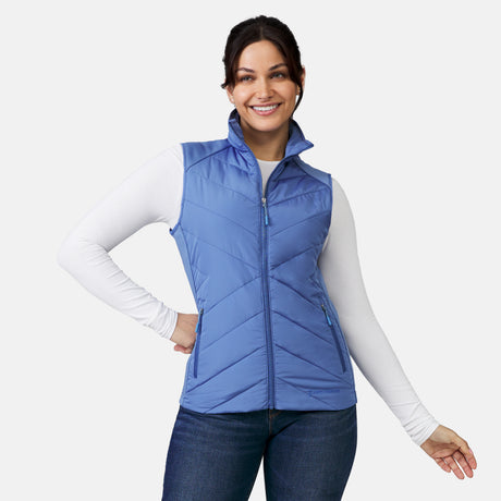 [Model is wearing a size Small.]  Woman wearing Free Country Women's Quilted Hybrid Vest in blue  with a full front zipper opening, patterned quilting, lower pocketse, and water resistant and wind resistant stretch performance fabric. light grey background. Perfect women’s jacket for winter weather.