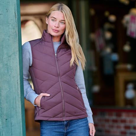 A woman wearing a plum-colored Women’s Quilted Hybrid Vest over a gray long-sleeved shirt and blue jeans.