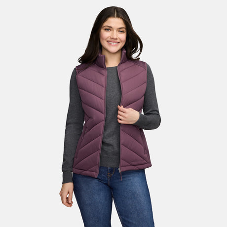 A woman wears a plum-colored Free Country Women’s Quilted Hybrid Vest with REPREVE synthetic fill over a grey long-sleeved shirt and blue jeans.