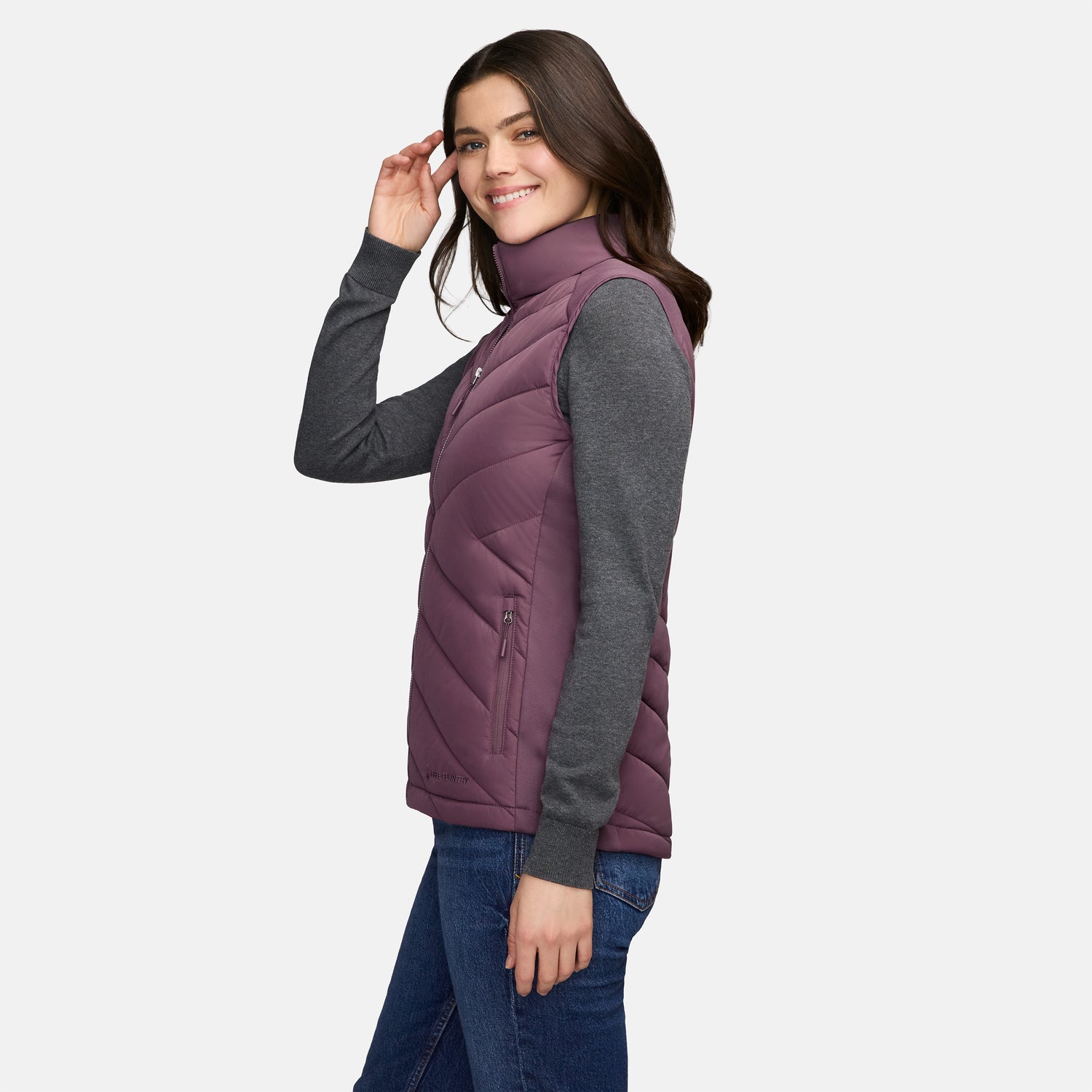 A woman wears a plum-colored Free Country Women’s Quilted Hybrid Vest over a grey long-sleeve shirt and blue jeans.
