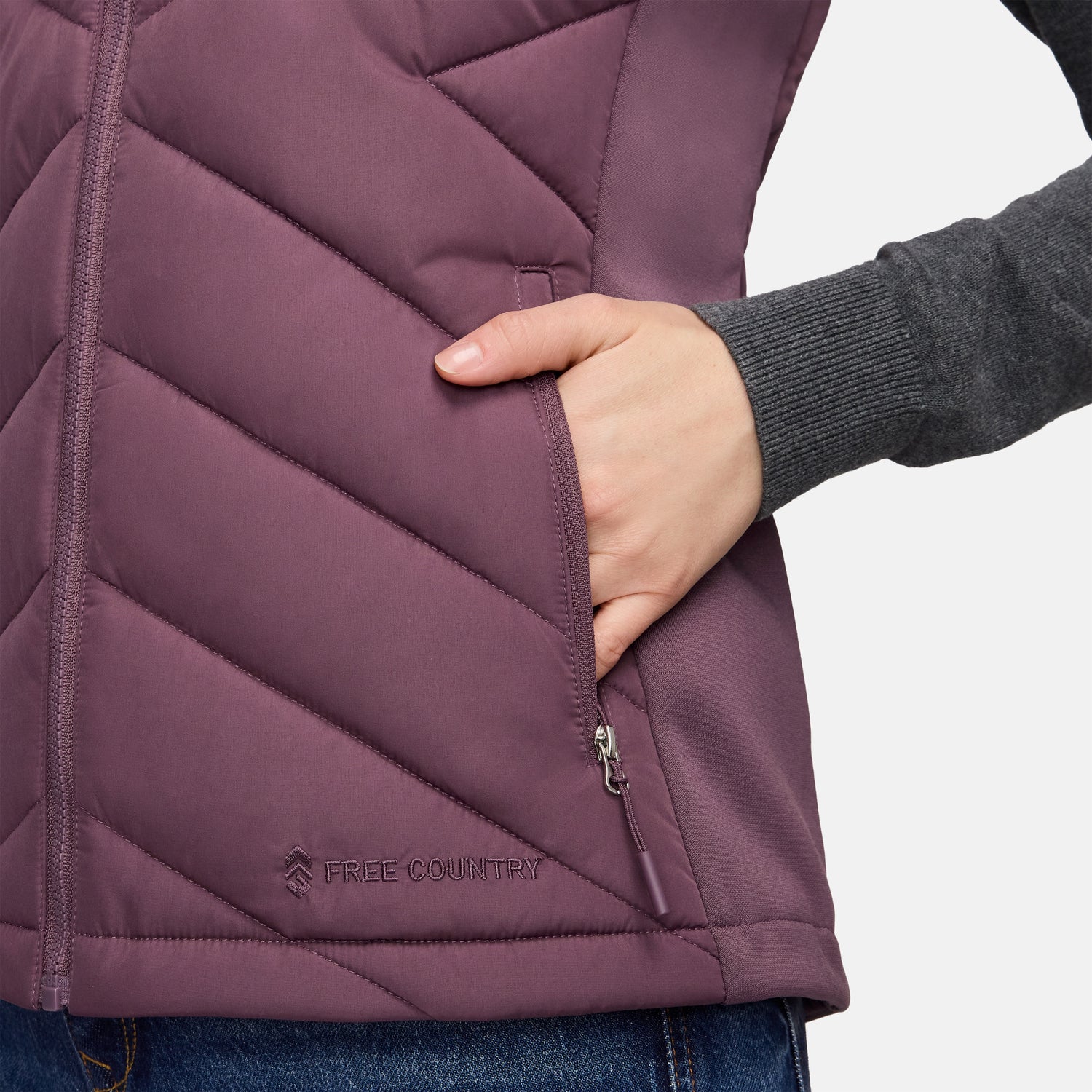 Close-up of a person wearing a plum-colored Free Country Women’s Quilted Hybrid Vest featuring a zippered pocket and a logo near the hem.