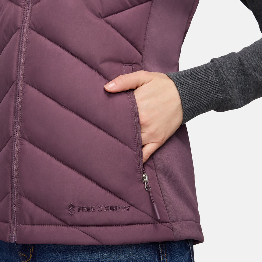 Close-up of a person wearing a plum-colored Free Country Women’s Quilted Hybrid Vest featuring a zippered pocket and a logo near the hem.