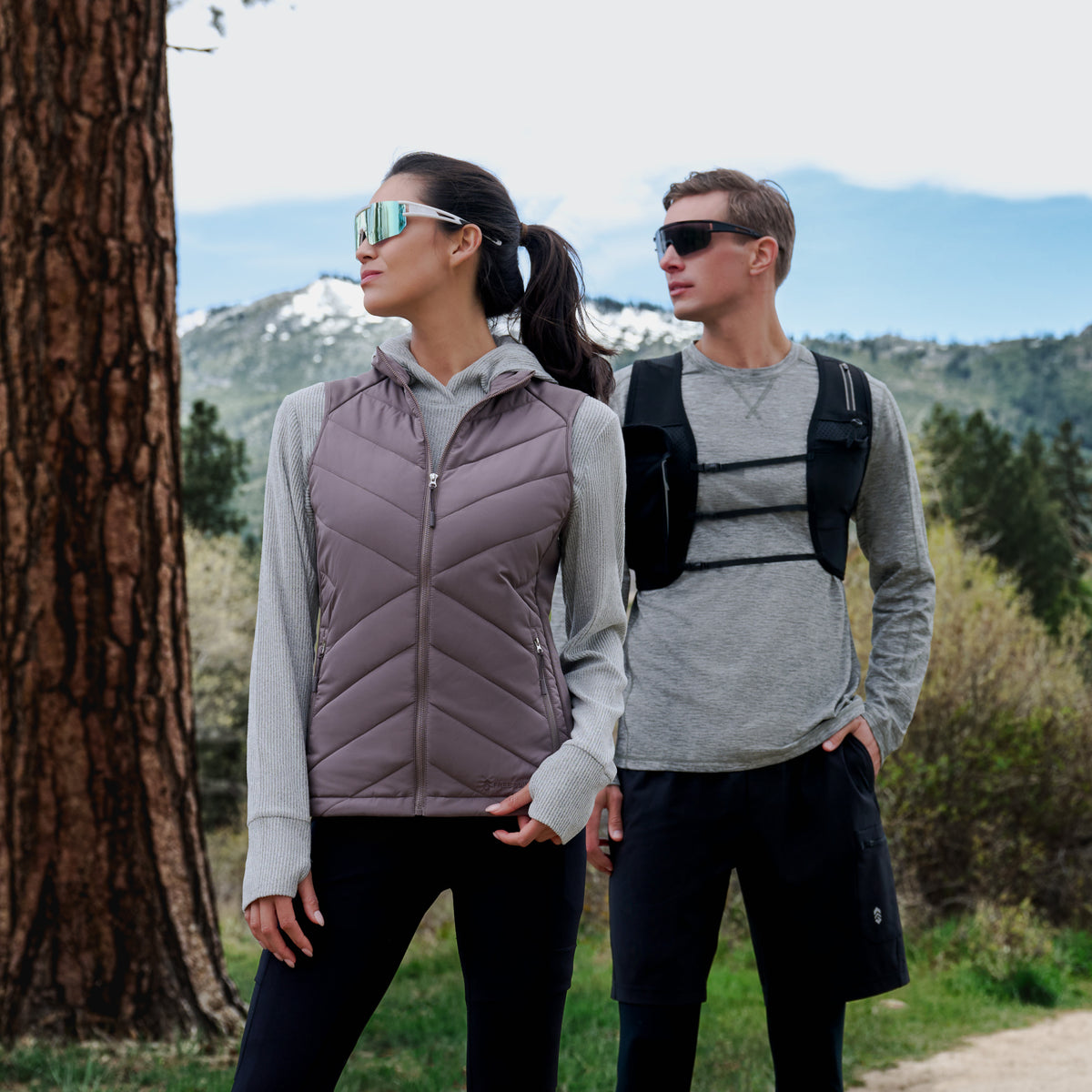 A man and a woman wearing matching gray hoodies and black athletic pants stand outdoors in a mountain setting. The woman wears a sleeveless quilted mauve-gray vest, and the man wears a black chest harness vest over his shoulders.