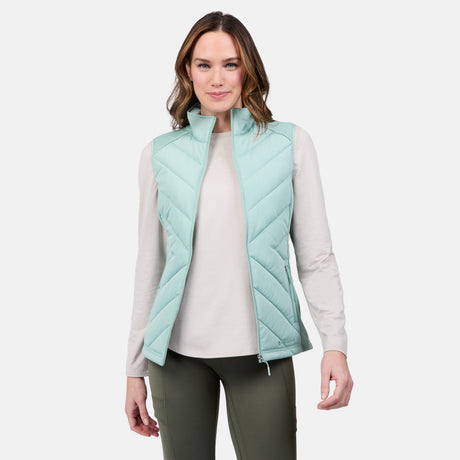 A woman with wavy brown hair models the Free Country Women's Quilted Hybrid Vest in light blue, featuring REPREVE synthetic fill, over a cream long-sleeve shirt and olive pants, smiling against a plain white background.