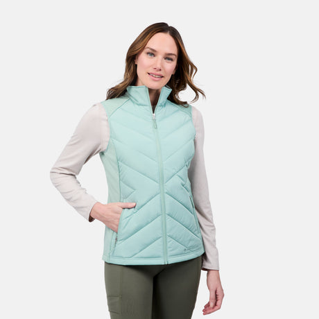 A woman models the Free Country Women's Quilted Hybrid Vest in light blue with REPREVE synthetic fill, layered over a beige long-sleeve shirt and green pants, standing with one hand in the vest pocket against a plain white background.