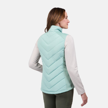 A woman faces away, wearing the Free Country Women's Quilted Hybrid Vest in light blue with REPREVE synthetic fill over a long-sleeve beige shirt and green pants, standing against a plain white background.