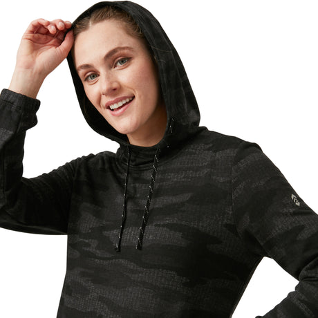 A smiling person wears the Women's Luxe Easy Fit Pullover by Clearance in a dark camo pattern, hood up and held with one hand, against a plain white background.