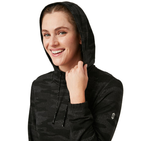 A smiling person wears the Clearance Women's Luxe Easy Fit Pullover in black camo with the hood up, holding the drawstring. The breathable hoodie has a small sleeve logo, set against a white background.
