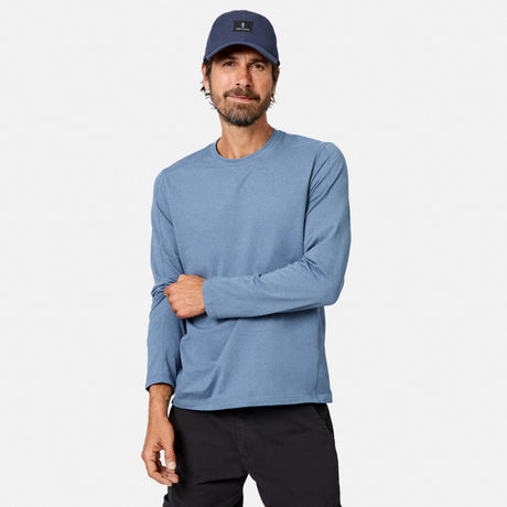 [Model is wearing size M.] A man wearing a heathered blue long-sleeve crewneck shirt, a navy blue baseball cap, and black pants, standing against a light gray studio background.