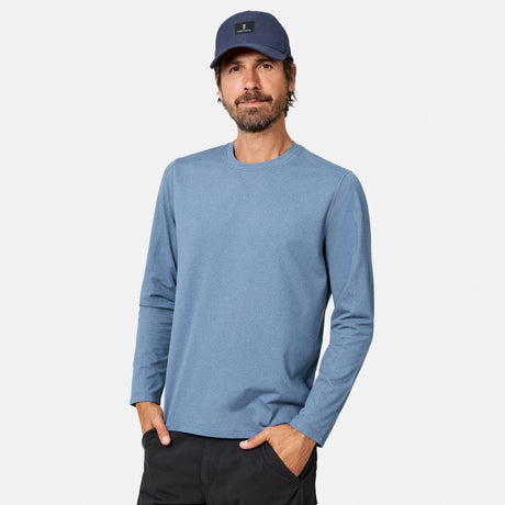 [Model is wearing size M.] A man wearing a heathered blue long-sleeve crewneck shirt, a navy blue baseball cap, and dark pants, standing against a light gray studio background.