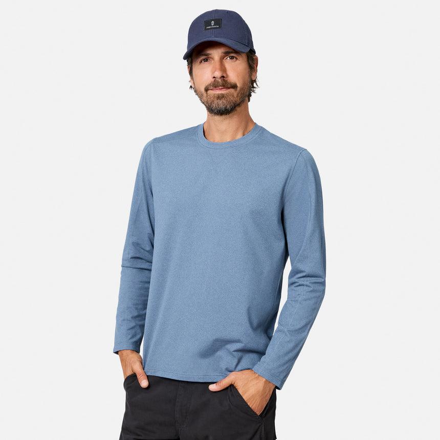 [Model is wearing size M.] A man wearing a heathered blue long-sleeve crewneck shirt, a navy blue baseball cap, and dark pants, standing against a light gray studio background.